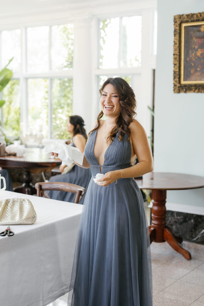 A bridesmaid laughs in front of large, brightly lit windows.