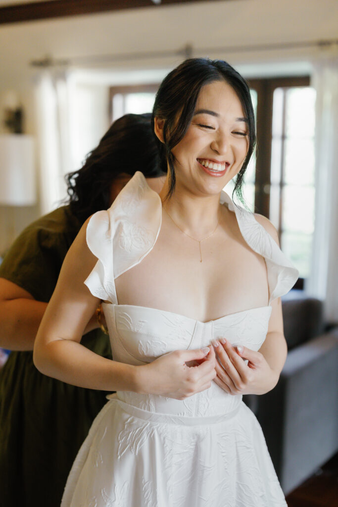 A bride laughs as her wedding dress is being fastened.