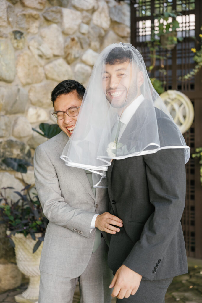 A groom laughs in surprise as his groomsman wears a veil. 